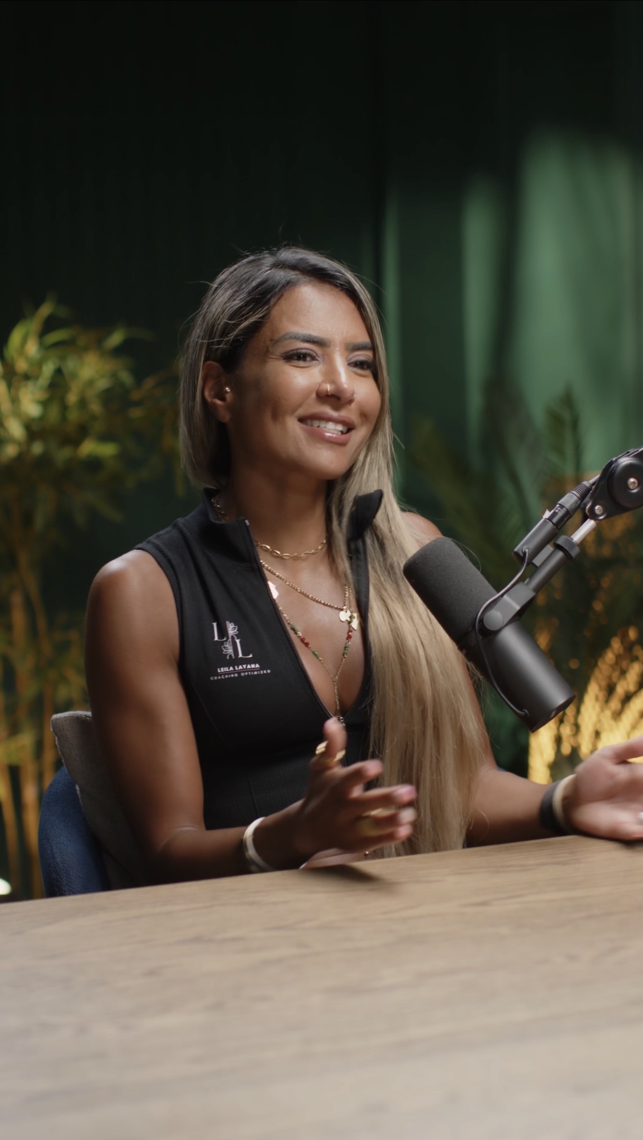 Leila Layana hosting in a Leila Layana Coaching Optimized branded zip-up tank, behind a podcast mic